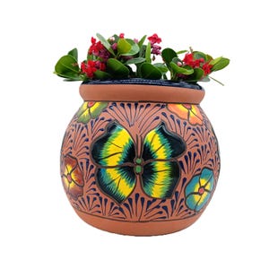 May include: A round terracotta planter featuring hand-painted floral motifs in yellow, green, blue, and orange against a natural clay background. The pot is filled with a small green plant with tiny red flowers, showcasing a dark blue rim.