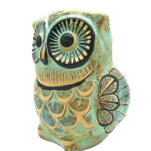 May include: A green ceramic owl figurine with gold and black painted details. The owl has large, round eyes with black pupils and gold accents. The body of the owl is covered in a pattern of scales, and the wings are decorated with a floral design.