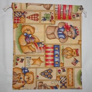 May include: A beige fabric drawstring bag with a patriotic theme. The bag is decorated with teddy bears holding American flags, stars, hearts, and the words "My Country, Sweet Land of Liberty, I Sing of."