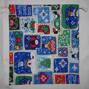 May include: A blue and white fabric with a Christmas theme. The fabric features a patchwork design with images of snowmen, candy canes, snowflakes, and the words "Let it snow" and "Caroling Cookies Cocoa".