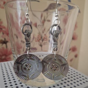 May include: A pair of dangle earrings featuring a silver-toned metal hand charm holding a circular ring, which is attached to a round, iridescent, gray-toned button with four central holes and a textured, radial pattern on the surface.