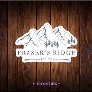 May include: A die-cut sticker featuring a blue ink-style illustration of mountain peaks and pine trees. The text reads FRASER'S RIDGE and EST. 1767 with arrow accents. Below the main design, the text nerdy bits is printed in a small purple font.