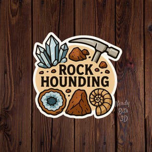 May include: A sticker with a beige background featuring the text "ROCK HOUNDING" and illustrations of crystals, a hammer, rocks, and a fossil. The sticker has a white border and is on a wooden background.