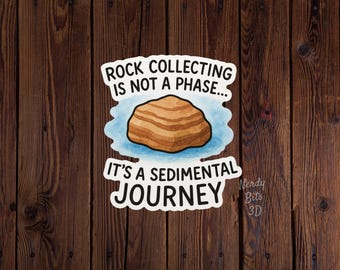 May include: A die-cut sticker featuring a brown, layered sedimentary rock illustration centered on a light blue watercolor-style background. The text reads: ROCK COLLECTING IS NOT A PHASE... IT'S A SEDIMENTAL JOURNEY. The sticker has a white border.