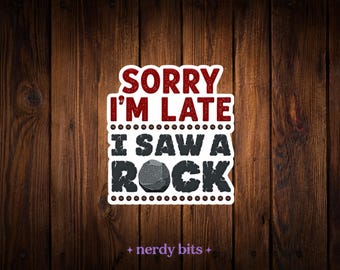 May include: A rectangular sticker with a white border featuring the red text SORRY I'M LATE and the dark gray text I SAW A ROCK, where the letter O in ROCK is replaced by a realistic illustration of a gray stone with a white highlight.