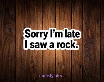 May include: A rectangular sticker with a white border and black text that reads Sorry I'm late I saw a rock. The sticker is centered against a dark brown vertical wooden plank background with the small purple text nerdy bits at the bottom.