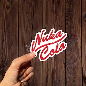 May include: A red and white Nuka Cola sticker with a distressed design. The sticker is held against a dark brown wooden background. The text "Nuka Cola" is in a stylized font.