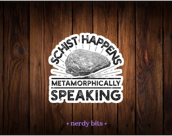 May include: A die-cut sticker featuring a gray rock illustration centered between the black text SCHIST HAPPENS and METAMORPHICALLY SPEAKING. The design has a distressed texture and a white border, with the small purple text nerdy bits below.