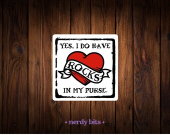 May include: A square white sticker with a black border featuring a red heart graphic wrapped in a banner that reads ROCKS. The text above the heart reads YES, I DO HAVE and the text below the heart reads IN MY PURSE. The sticker has a distressed edge.