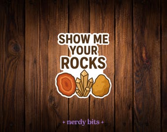 May include: A die-cut sticker featuring the brown text SHOW ME YOUR ROCKS above illustrations of a red-orange agate slice, a cluster of tan crystals, and a brown rock. The sticker has a white border and is centered on a dark wood background.
