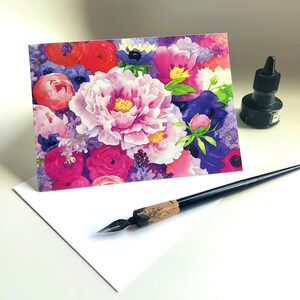 May include: A greeting card featuring a watercolor floral design with pink peonies, red poppies, and purple accents. A white envelope, black ink bottle, and a calligraphy pen are also visible.