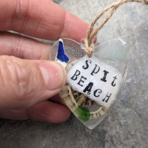 May include: A heart-shaped glass ornament with a blue and white seashell, green glass pieces, and the text "Spit Beach" inside. The ornament is hanging from a twine string.