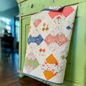 Puede incluir: Una colcha de retazos con fondo blanco y patrones coloridos en forma de lazo en tonos azules, rosas, verdes y naranjas. La colcha está doblada y apoyada en una cómoda verde.