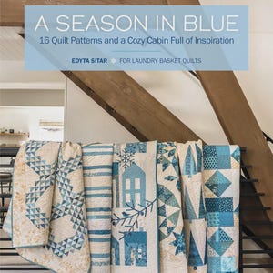 May include: A display of several blue and white quilts, each with unique patterns, including geometric shapes and house motifs. The quilts are hanging on a black metal rack. The image also features a book cover that reads "A Season in Blue."