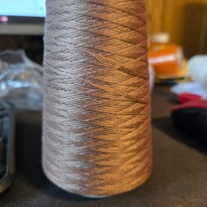 May include: A large spool of metallic bronze-colored thread wound in a tight, crisscross pattern. The cylindrical spool features a smooth, light-colored plastic core at the top, with the thread covering the entire body in a consistent, dense weave.