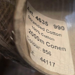 May include: A spool of 100% cotton thread with a white label reading Art. 4635 990 Stranded Cotton 100% Cotton 2000m Conen Colour: 856 Lot: 44117. The thread is wrapped around a cylindrical cardboard core and partially covered in clear plastic film.
