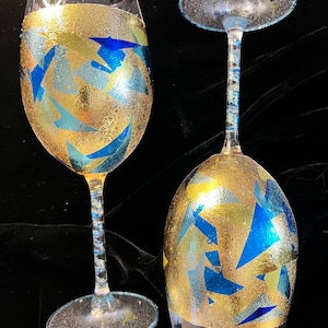 May include: Two clear glass wine glasses with gold and blue geometric patterns. The glasses have a sparkly finish.