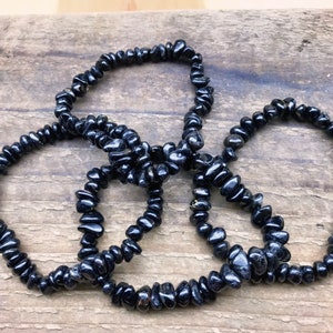 May include: Four black gemstone chip bracelets, made of small, irregular shaped beads, are stacked on a wooden surface.