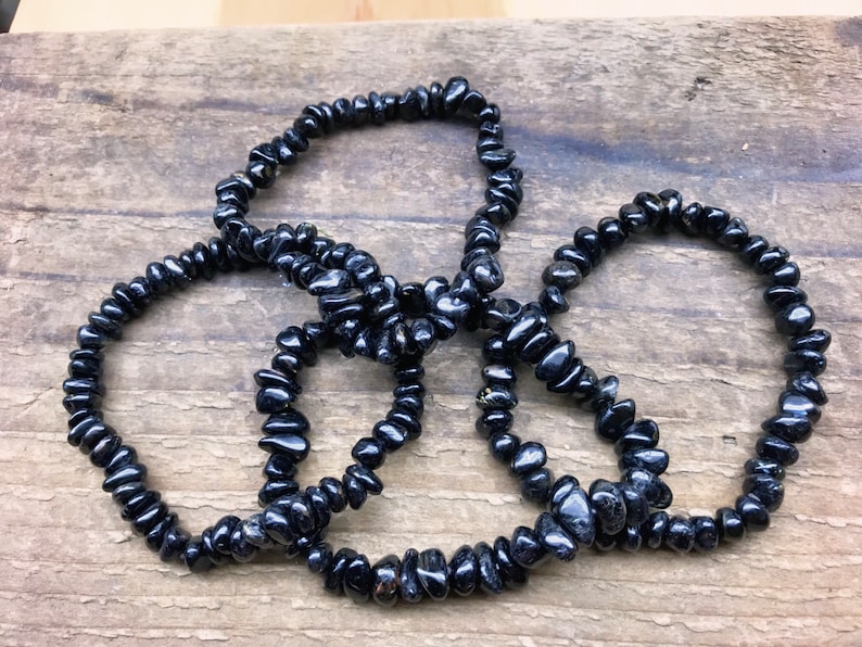 May include: Four black gemstone chip bracelets, made of small, irregular shaped beads, are stacked on a wooden surface.