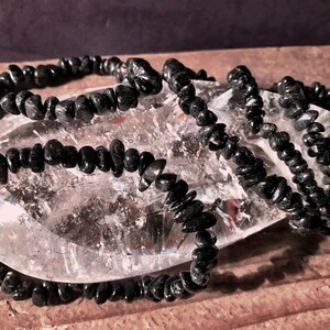 May include: A strand of black gemstone chips, likely obsidian, arranged in a loose pile on a clear quartz crystal. The chips are small and irregular in shape, creating a textured look.