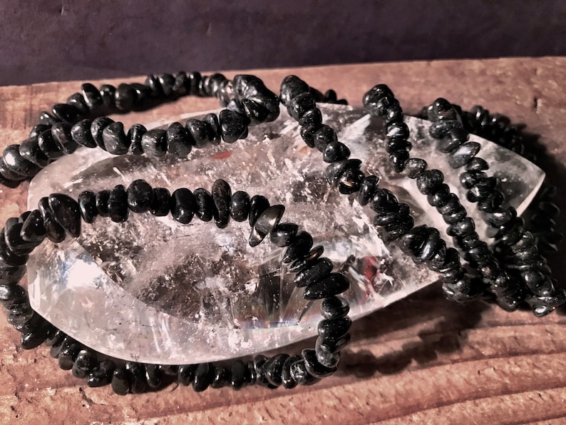May include: A strand of black gemstone chips, likely obsidian, arranged in a loose pile on a clear quartz crystal. The chips are small and irregular in shape, creating a textured look.