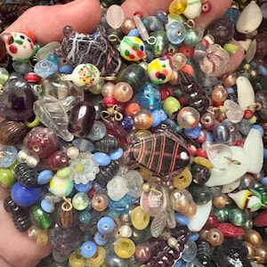 May include: A collection of colorful glass beads in various shapes and sizes, including round, floral, and animal-shaped beads. The beads are in shades of blue, green, red, yellow, and clear, with some featuring intricate designs and patterns. The beads are held in a hand.