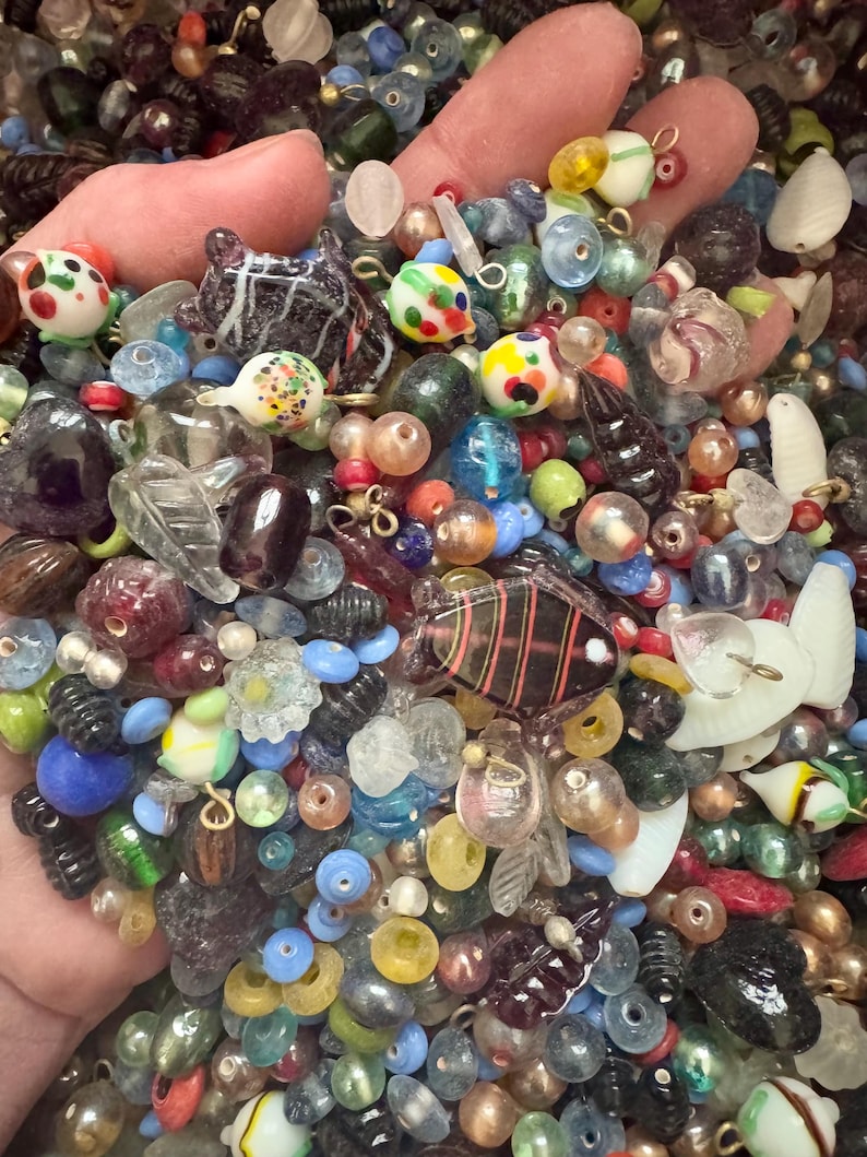 May include: A collection of colorful glass beads in various shapes and sizes, including round, floral, and animal-shaped beads. The beads are in shades of blue, green, red, yellow, and clear, with some featuring intricate designs and patterns. The beads are held in a hand.