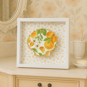 May include: A framed artwork featuring a seashell design with oranges, leaves, and white flowers. The artwork is set against a floral patterned background and framed in a white square frame. The seashell has a gold trim.