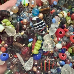 May include: A collection of colorful glass beads in various shapes and sizes. The beads include round, oval, and irregular forms, with colors like red, blue, green, and clear. Some beads have striped or patterned designs, and some have metal loops.