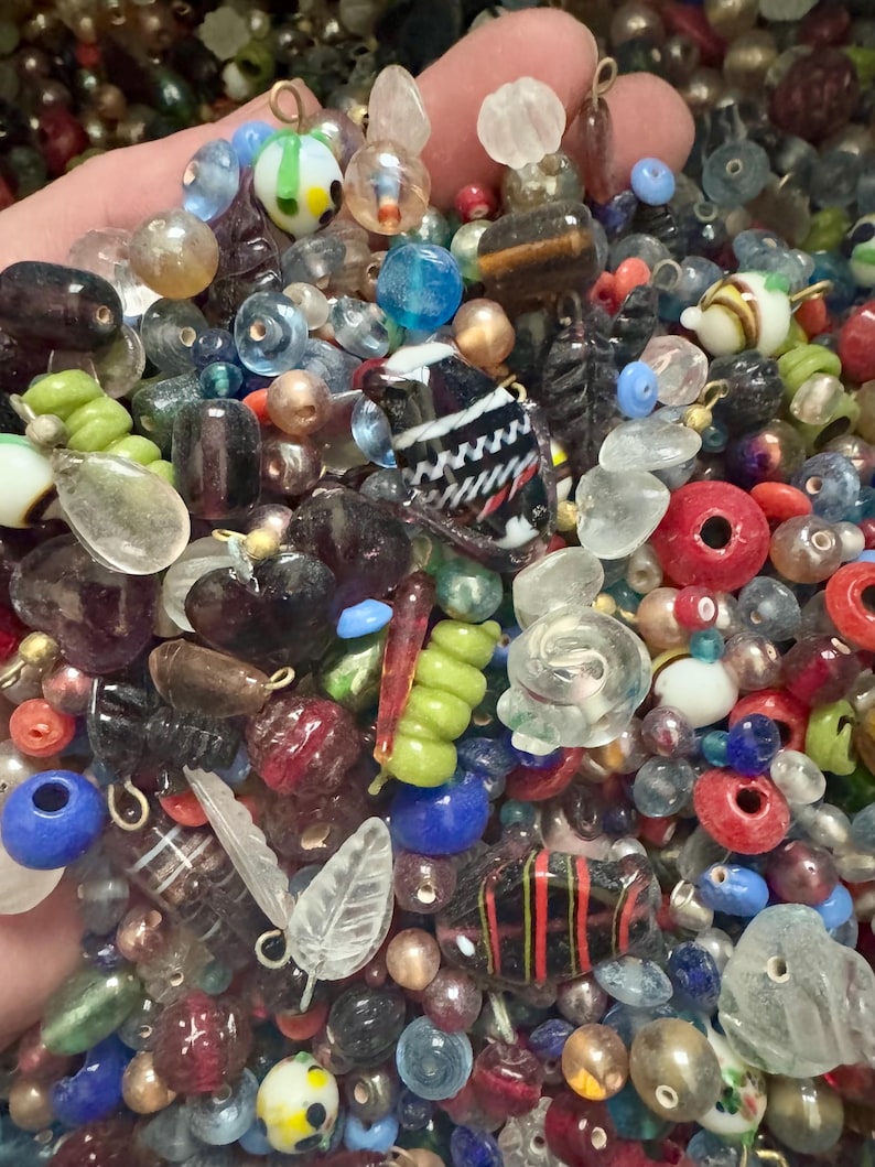 May include: A collection of colorful glass beads in various shapes and sizes. The beads include round, oval, and irregular forms, with colors like red, blue, green, and clear. Some beads have striped or patterned designs, and some have metal loops.