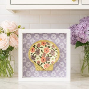 May include: A framed seashell art piece. The seashell is filled with a floral pattern in shades of pink, peach, and purple. The shell is outlined with gold glitter and set against a lavender background with white flower shapes. The frame is white.