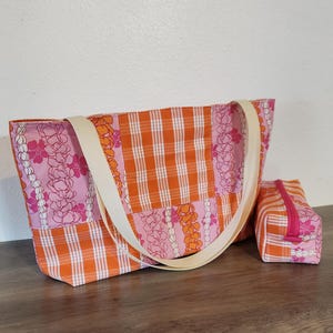 May include: A tote bag and a matching rectangular zippered pouch featuring a patchwork design of orange and white gingham fabric alongside pink fabric printed with orange and pink floral lei patterns, finished with two cream-colored fabric straps.