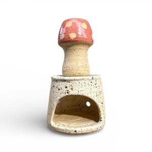 May include: A ceramic mushroom-shaped oil warmer. The mushroom cap is red with white spots, and the stem is beige. The base is a speckled beige with a large opening and a small hole. The warmer is designed to hold essential oils.