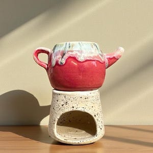 May include: A ceramic teapot with a red body, white and blue accents, and a small spout. It sits atop a speckled, off-white ceramic base with a rounded opening. The teapot has a handle and is placed on a wooden surface.