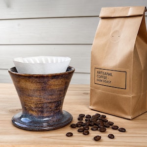 May include: A ceramic coffee dripper with a mottled brown and blue glaze containing a white paper filter, positioned next to a brown paper bag labeled "ARTISANAL COFFEE - DARK ROAST" and a small pile of dark roasted coffee beans.