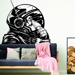May include: Black and white illustration of a monkey wearing a space helmet, looking thoughtful. The illustration is on a white wall above a dark blue couch with pink and gray throw pillows.