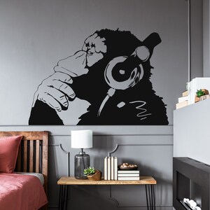 May include: Black and white silhouette wall decal of a gorilla wearing headphones. The gorilla is looking to the side and has its hand on its chin, as if in thought. The decal is on a gray wall in a room with a wooden bed, a side table, and a fireplace.