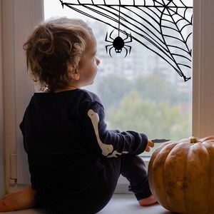 Puede incluir: Un niño vestido con un disfraz de esqueleto negro se sienta junto a una ventana decorada con una telaraña y una araña. Una gran calabaza naranja se encuentra en el alféizar de la ventana junto a una vela blanca y una linterna negra.
