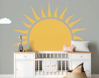 May include: A white crib with a white dresser and a yellow sun decal on the wall. The crib has a white mattress and a white blanket with a gray pattern. The dresser has two drawers and a white top. The sun decal is on the wall behind the crib and dresser.