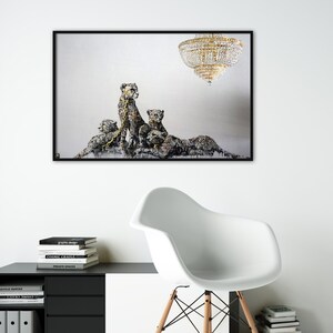May include: A framed print of a painting depicting four cheetahs in a white room with a large crystal chandelier hanging from the ceiling. The cheetahs are painted in a realistic style with a focus on their fur and spots. The painting is framed in a black frame.