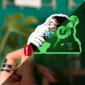 May include: A green glow-in-the-dark sticker of a gorilla wearing headphones. The gorilla's face is covered in colorful paint splatters.