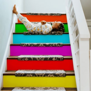 May include: A child is lying on a staircase with colourful painted steps. The steps are painted in a rainbow pattern, starting with red at the top and ending with yellow at the bottom. The child is wearing a white and yellow patterned shirt and has their hair tied back.