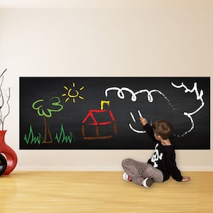 May include: A child draws on a black chalkboard wall decal with colorful chalk. The decal is on a white wall and features a house, a tree, grass, and a sun.