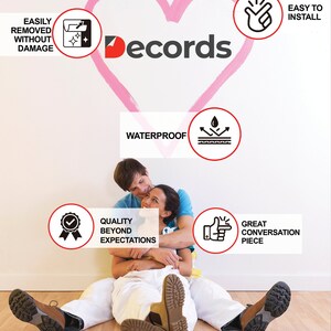 May include: A couple sits on a wooden floor in front of a white wall with a pink heart painted on it. The heart has the word "Decords" written on it. There are several red circles with white icons and text inside them. The text includes "Easily Removed Without Damage", "Waterproof", "Easy to Install", "Quality Beyond Expectations", and "Great Conversation Piece".