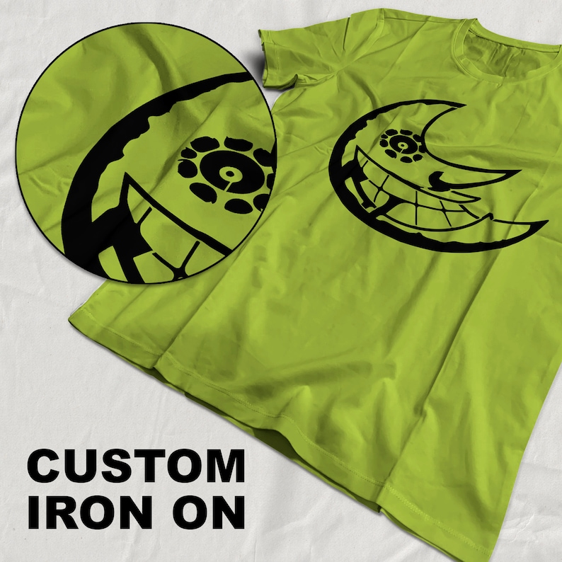 May include: A green t-shirt with a black iron-on design of a crescent moon with a face and a circle in the center. The text "CUSTOM IRON ON" is printed below the image.