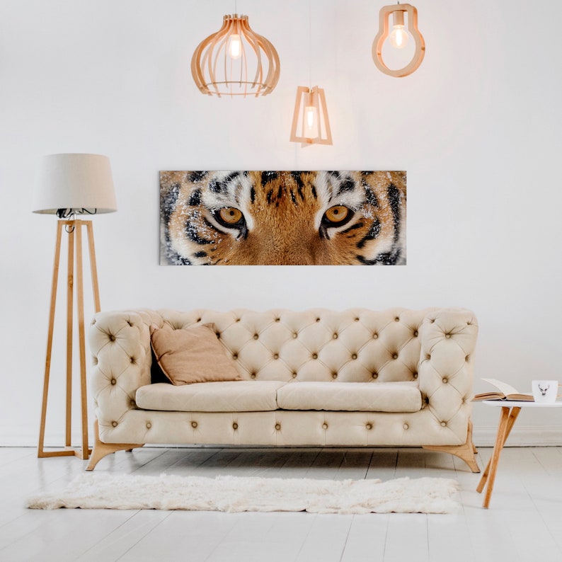 May include: A close-up of a tiger's face with orange and black stripes. The tiger's eyes are open and staring directly at the camera. The tiger's face is in focus, while the background is blurred. The image is printed on a canvas and hung on a white wall above a white couch.