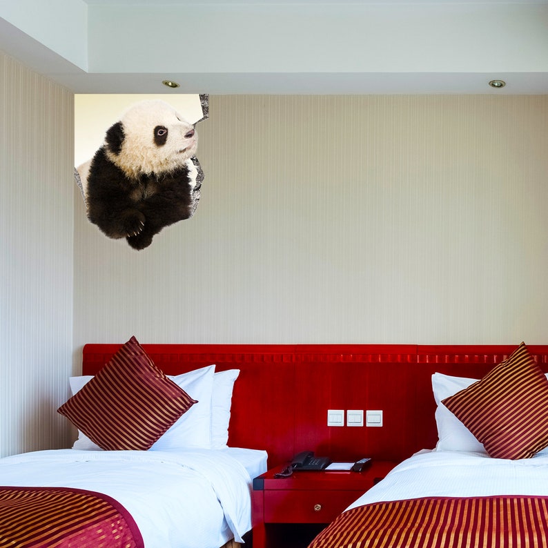 May include: A black and white panda bear peeking out of a hole in a white ceiling above a red headboard with two twin beds. The beds have white sheets and red and gold striped bedspreads.