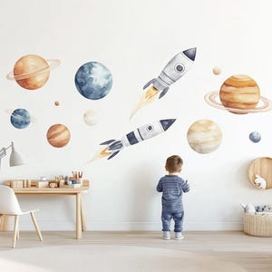 Può includere: Un muro bianco con un adesivo murale a tema spaziale che presenta pianeti, stelle e razzi. I pianeti sono in tonalità di marrone e blu, e i razzi sono bianchi e grigi con fiamme arancioni. L'adesivo è perfetto per la camera da letto di un bambino o una sala giochi.