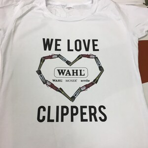 May include: White t-shirt with the text "WE LOVE" above a heart shape made of clippers. The heart shape includes the text "WAHL" and the brands "WAHL", "MOSER", and "ermila". The text "CLIPPERS" is below the heart.