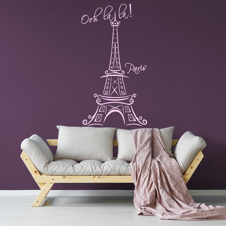 May include: A white outline of the Eiffel Tower with the text "Ooh la la!" and "Paris" on a purple wall. A light wood couch with white cushions and a pink throw blanket is in front of the wall.
